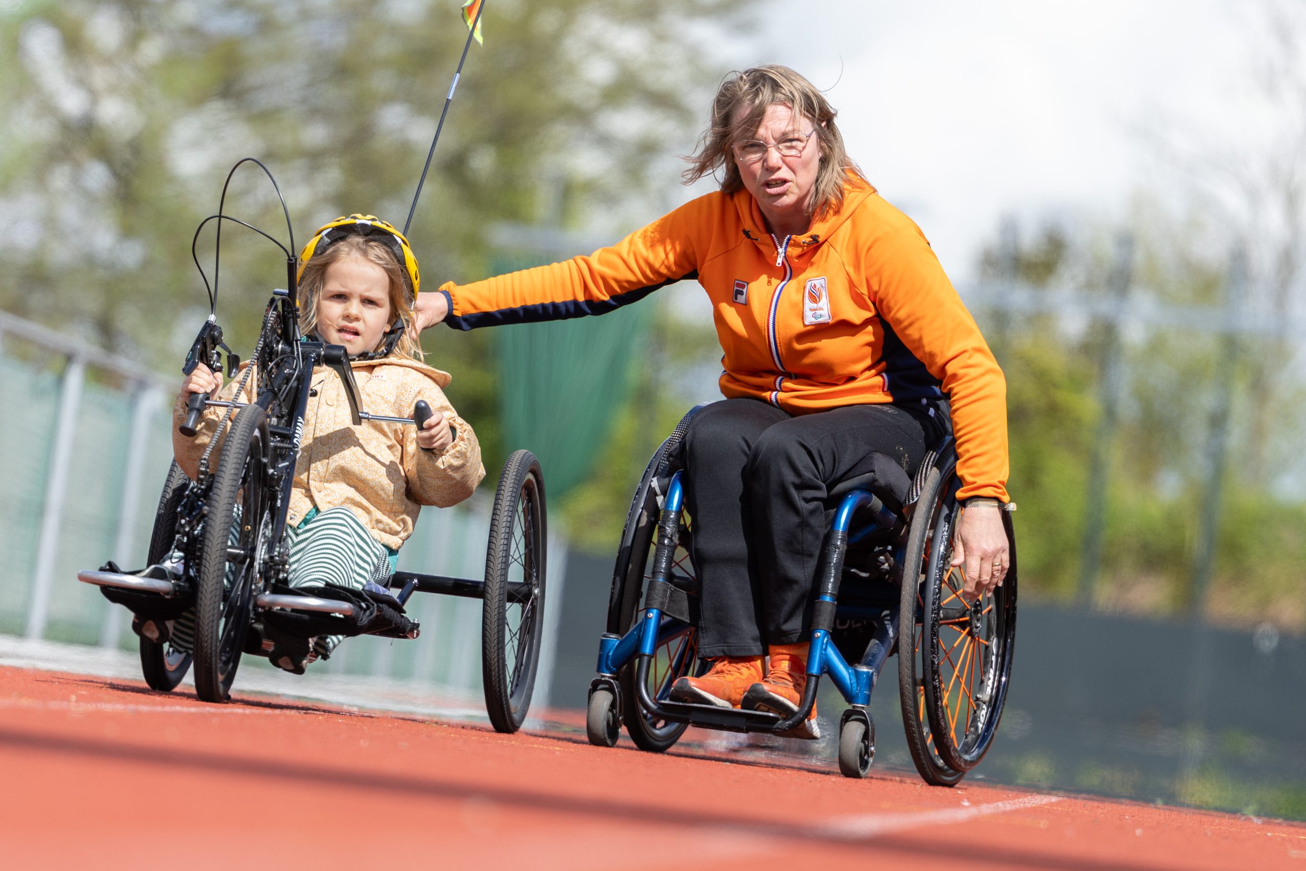 Para-triathleet Margret IJdema rijdt in een handbike naast een jonge deelnemer van een handbike evenement. Ze rijden op een atletiekbaan en Margret begeleidt het kind.