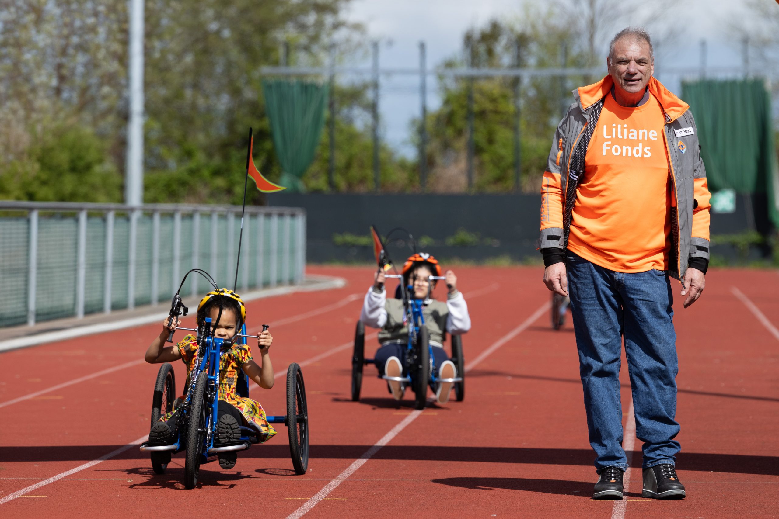 Twee kinderen rijden met een handbike over een atletiekbaan, begeleid door actievoerder Alessandro. Hij zet zich in voor het Liliane Fonds en organiseert het handbike‑evenement