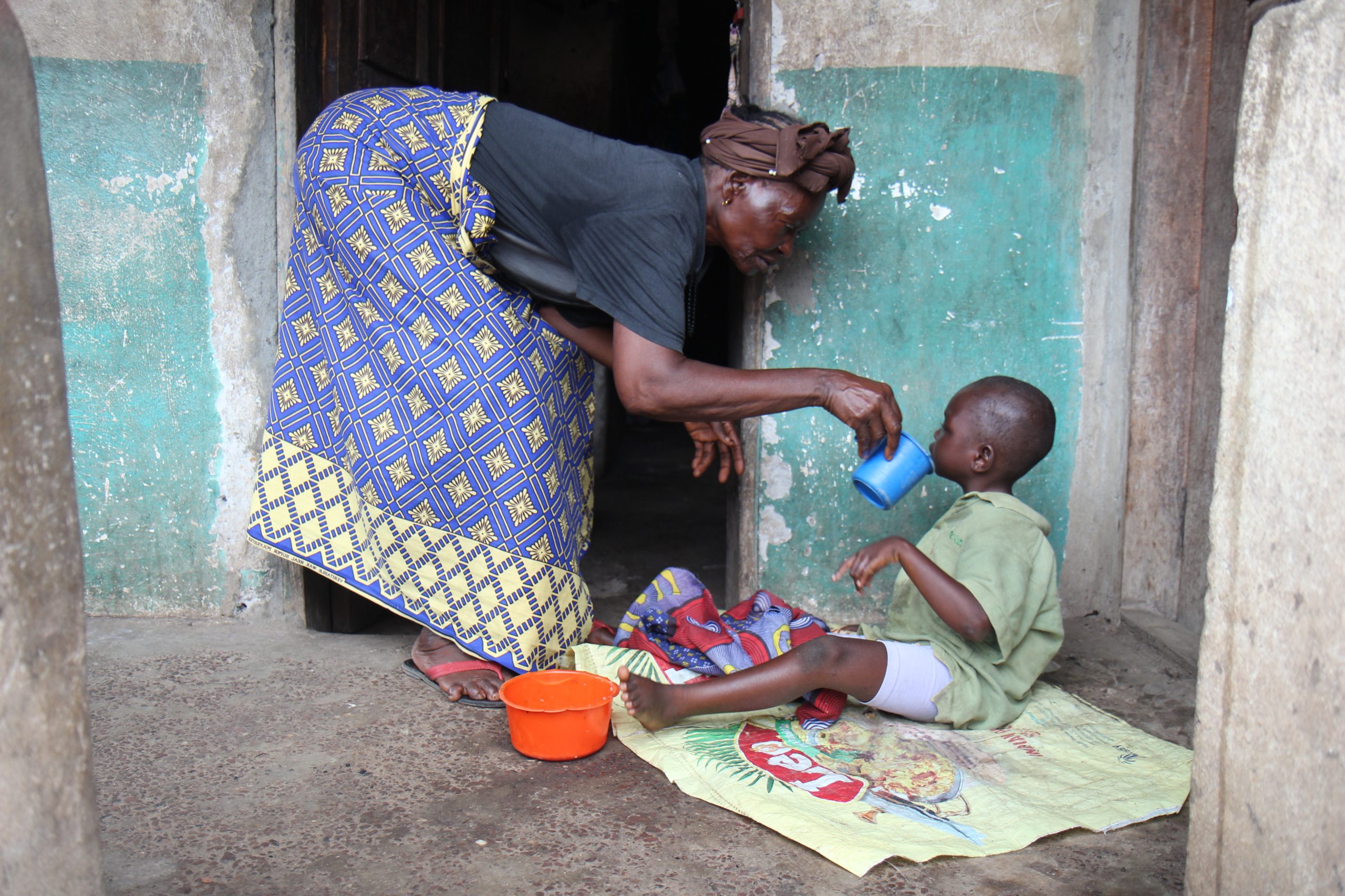 Oma Reba en haar kleinzoon Alhaji zitten voor hun huis in Sierra Leone. Alhaji zit op een kleedje terwijl zijn oma hem wat te drinken geeft